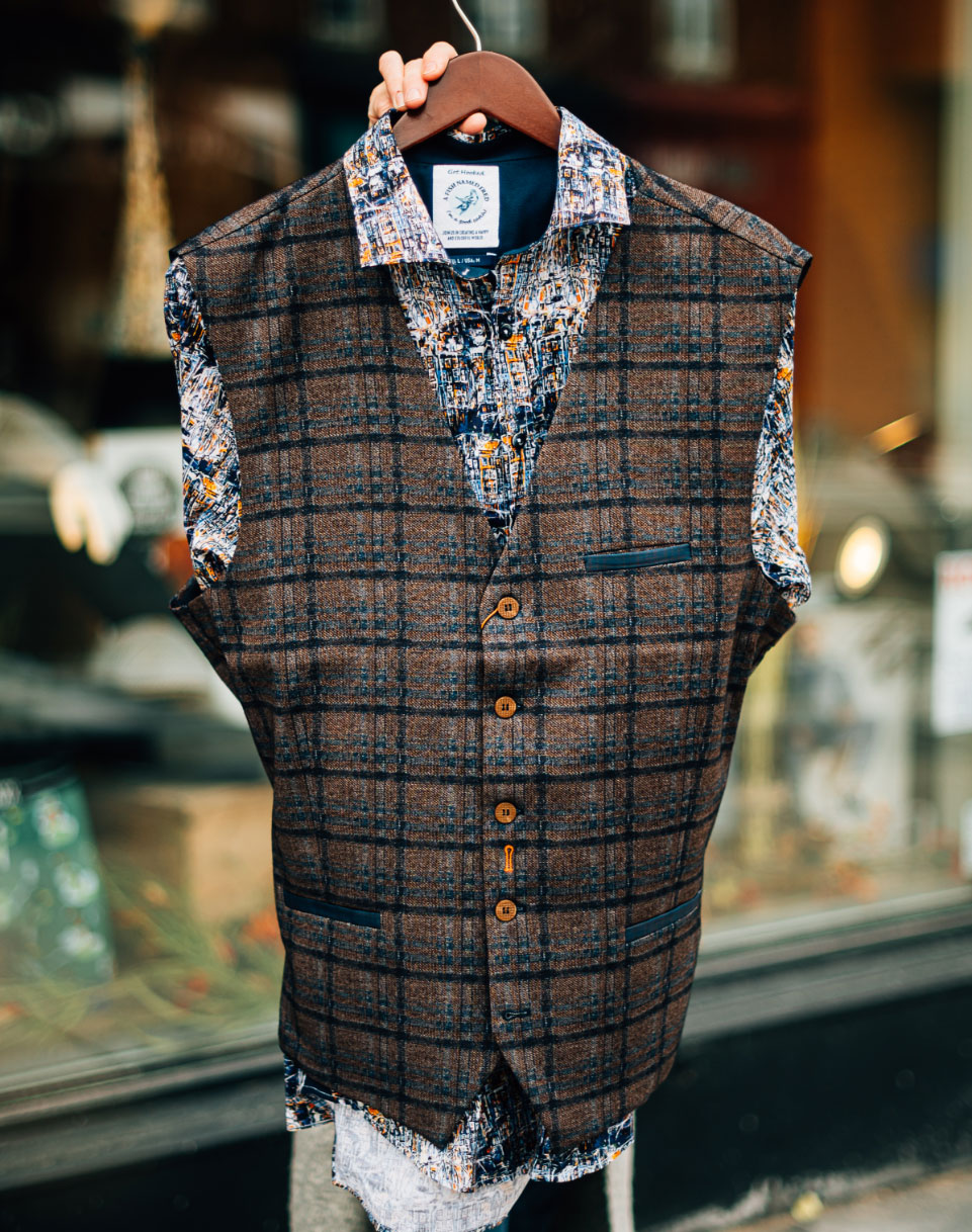 A brown and blue plaid men’s vest over top a colourful dress shirt on a coat hanger